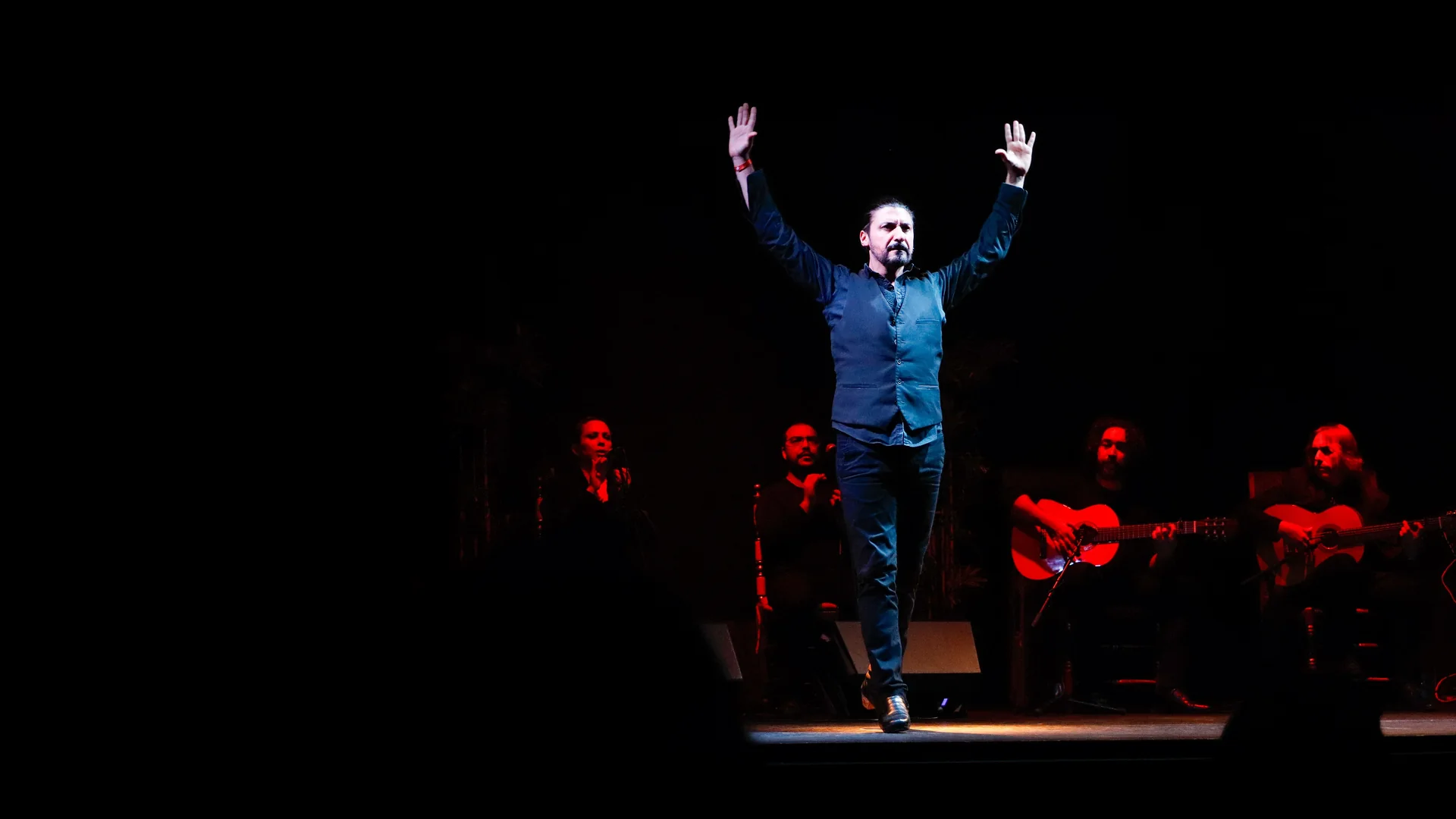 A flamenco dancer in a dark blue waistcoat stands with both arms raised under a single spotlight, with a clapping singer and two guitarists seated behind him on a dark stage