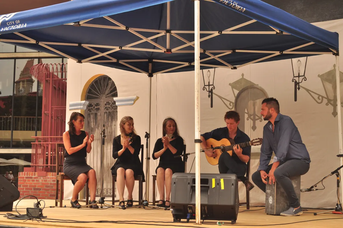 Three women clapping palmas beside a guitarist and a cajón player on an outdoor flamenco stage