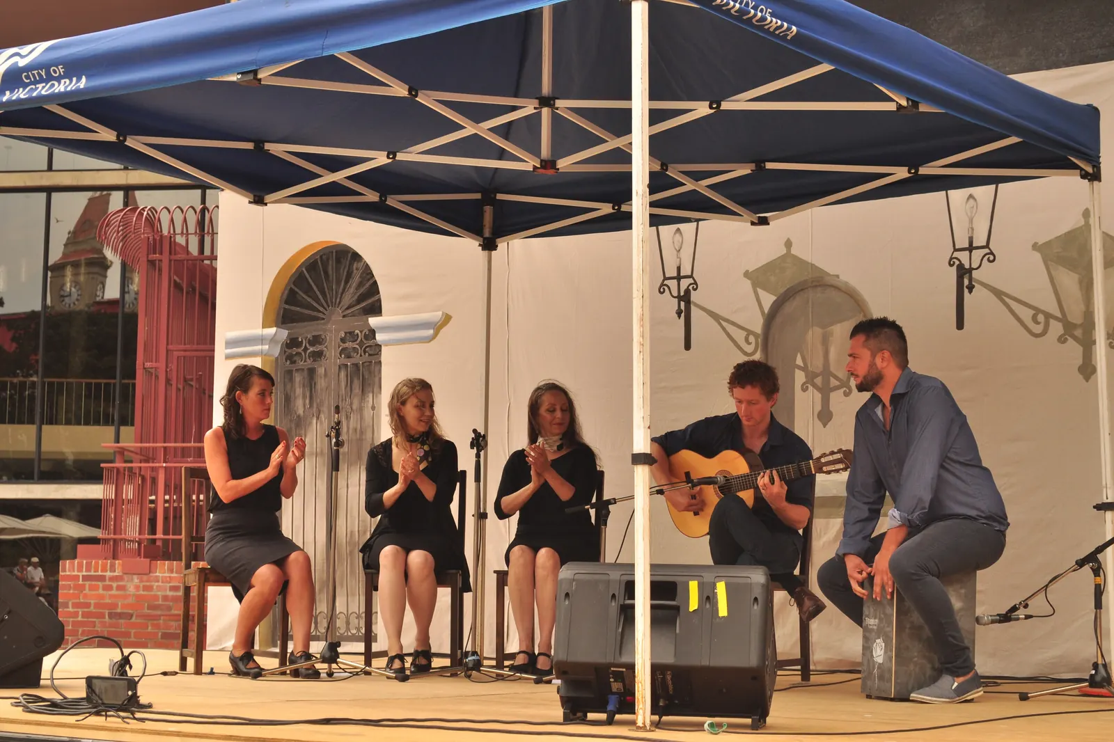 Three women clapping palmas beside a guitarist and a cajón player on an outdoor flamenco stage