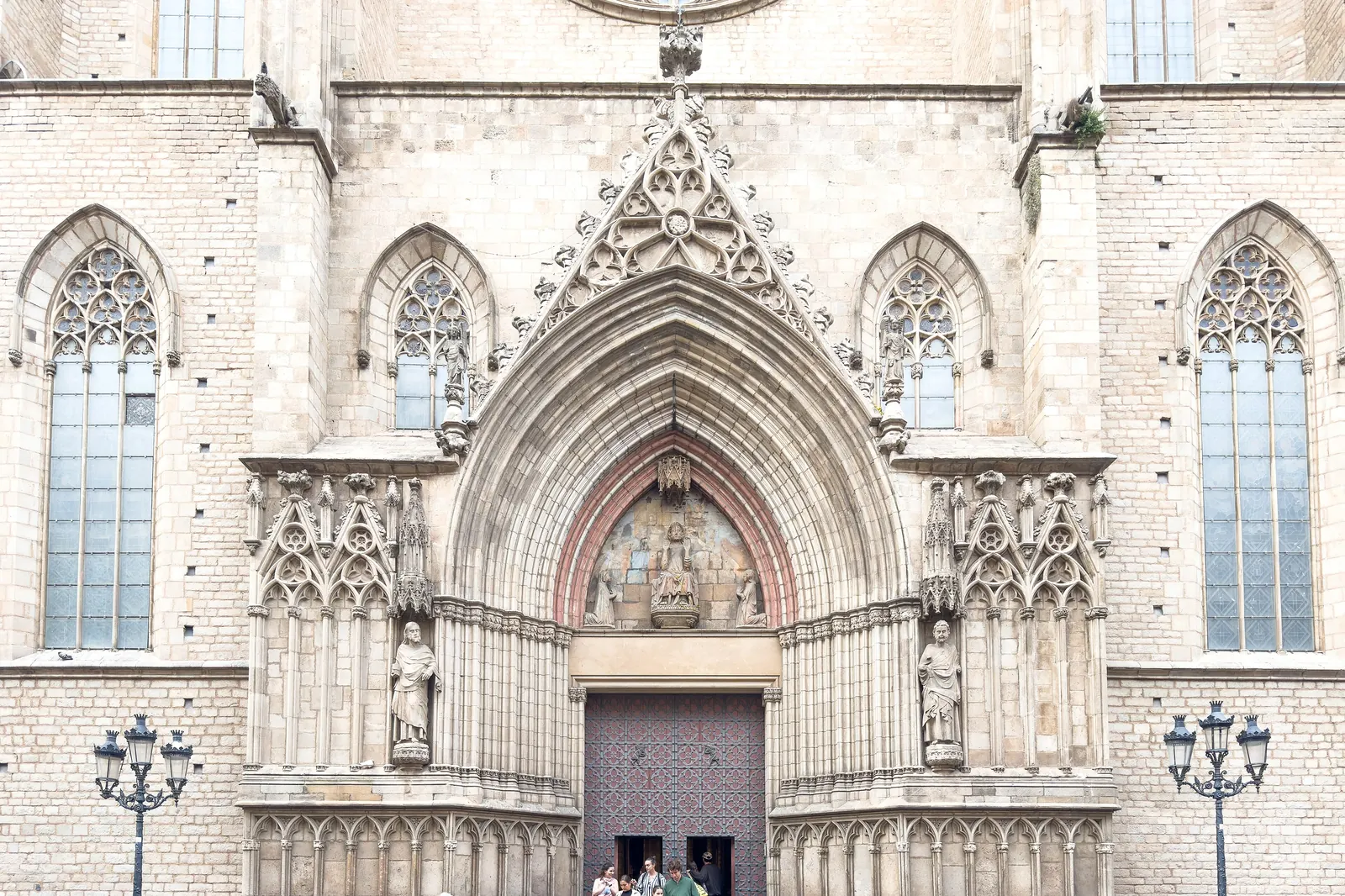 The west portal of Santa Maria del Mar, a pointed Gothic arch with carved statues, a dark wooden door and visitors standing on the steps